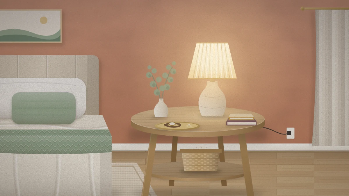 Small round wooden nightstand with a lit ceramic lamp, a bud vase of eucalyptus, a brass tray holding a watch and ring, and two stacked books