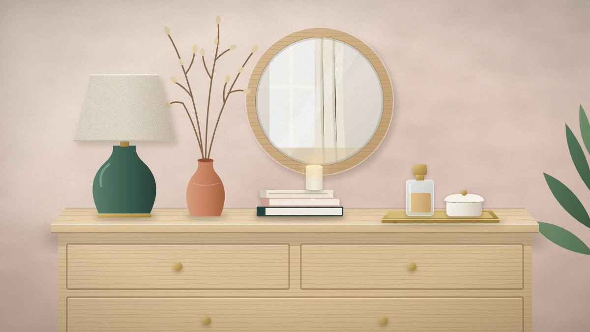 Pale oak dresser top with a round wood-framed mirror above, a cream-shaded lamp, a tray with a perfume bottle and box, books and tall dried branches